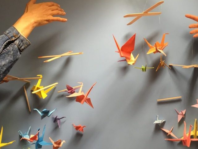 Hands holding up four finished colourful origami paper crane mobiles.